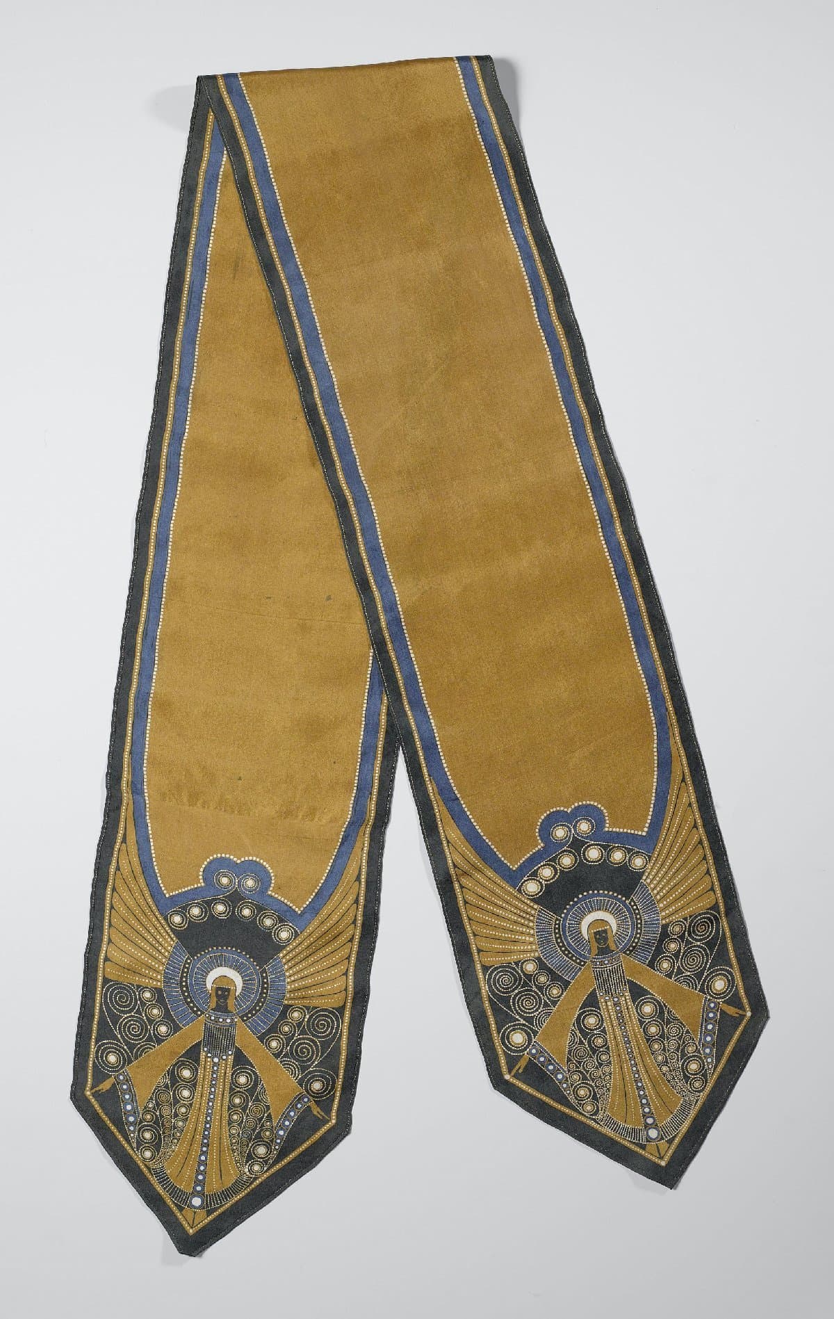 Langwerpige gebatikte sjaal in geel-goud, blauw, wit en groen, met onbewerkt middenstuk en aan beide uiteinden een staande figuur met gestrekte gespreide armen